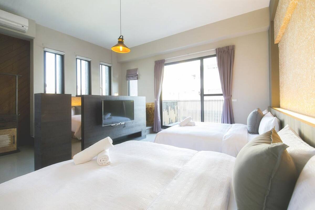 Habitación de hotel con dos camas, televisión, escritorio y balcón con cortinas.