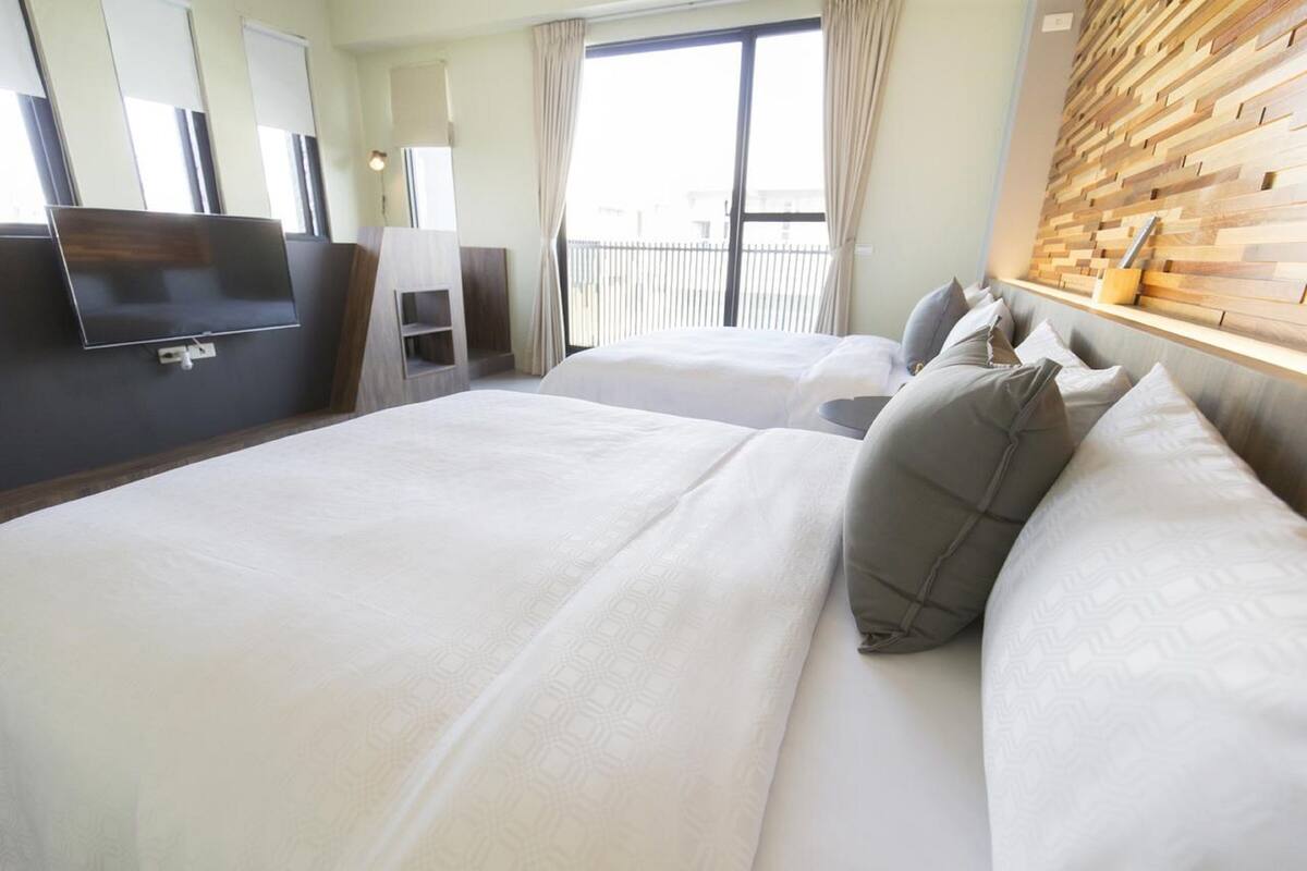 Habitación de hotel con dos camas, un televisor montado en la pared, cabecera de madera y un balcón con cortinas.