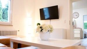 A modern living room with a white sofa, a white coffee table, a flat-screen TV mounted on the wall, a small potted plant, and a window with a view of greenery. - Ferienhaus Domblick Herkenrath (Bergisch Gladbach)