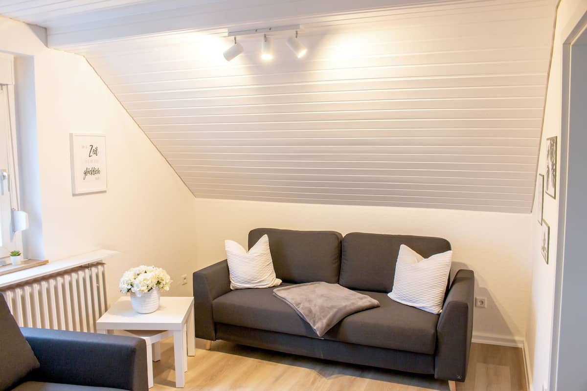 A modern living room with a dark grey sofa, white cushions, a small white table with a vase of flowers, and a white radiator.