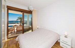 Un dormitorio con una cama grande, una mesita de noche, una lámpara y vistas a una piscina y palmeras a través de un ventanal.