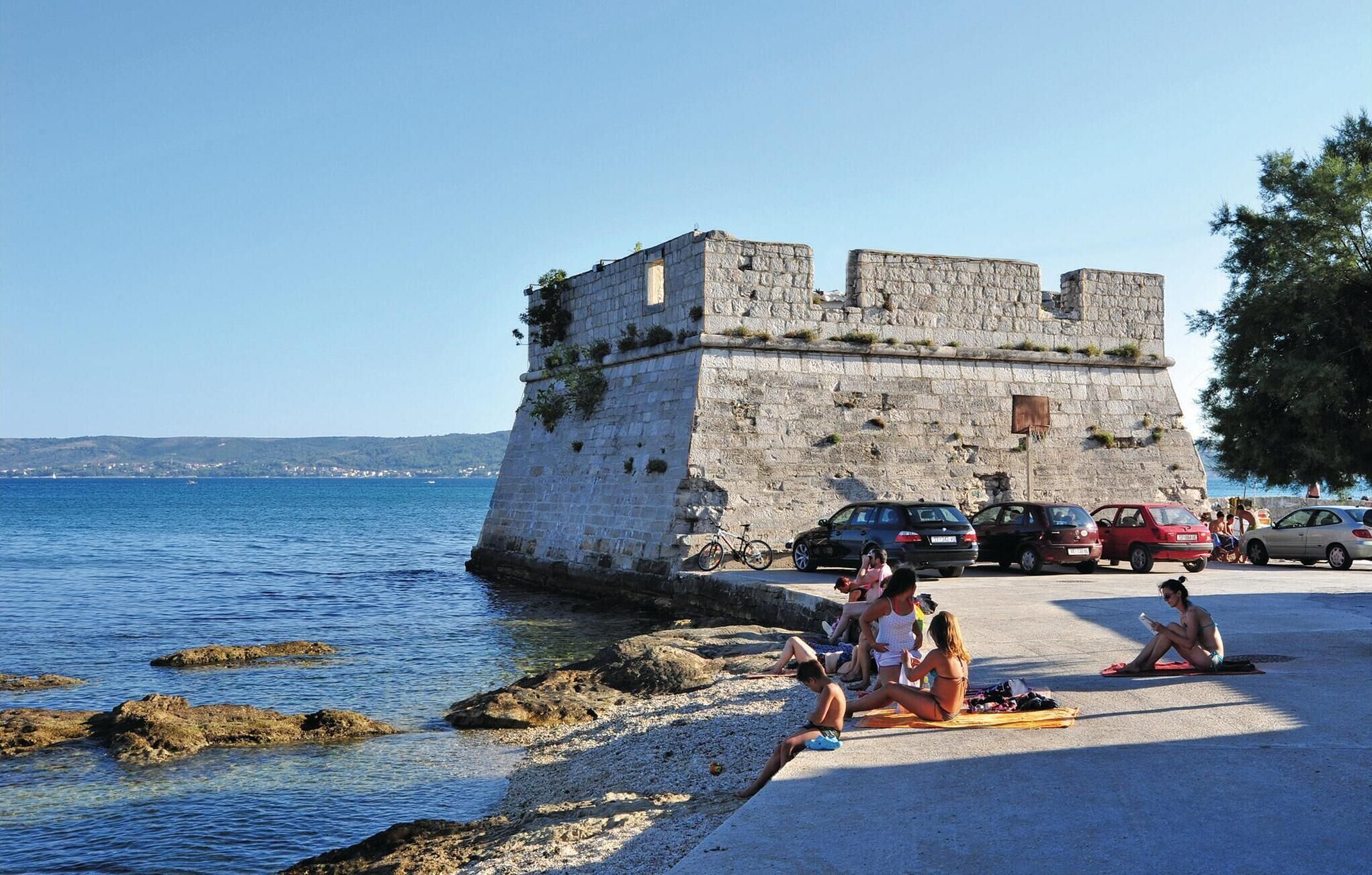A coastal area with a stone fortification, a beach with people sunbathing, and parked cars along the shore.