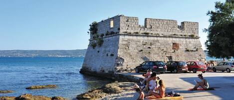 A coastal area with a stone fortification, a beach with people sunbathing, and parked cars along the shore.