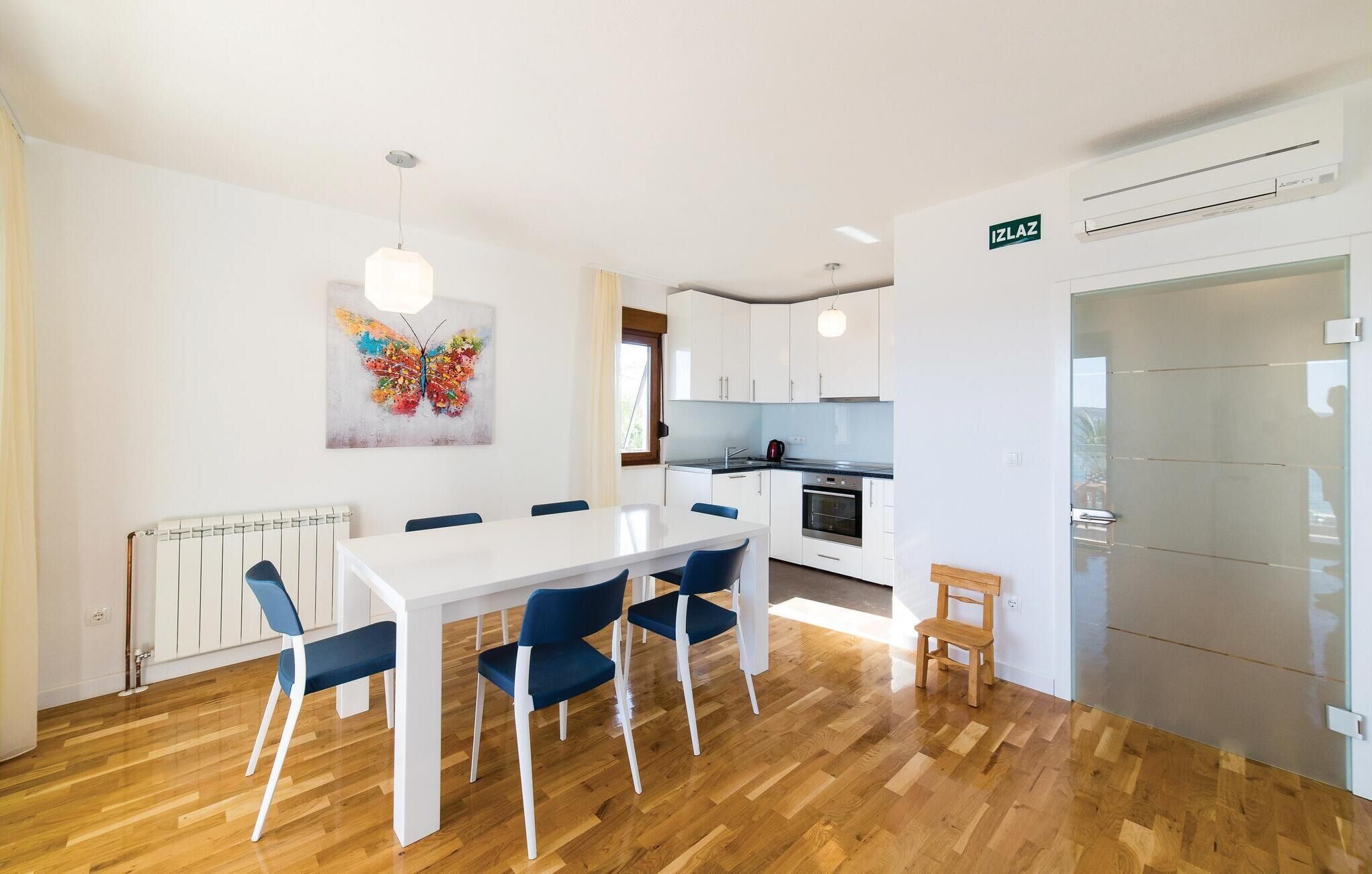 A modern kitchen with white cabinets, a stove, and a sink. There is a dining area with a white table and blue chairs. A colorful butterfly painting hangs on the wall.