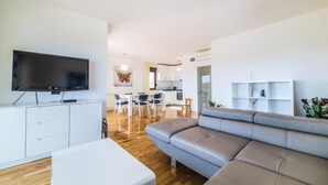 Een moderne woonkamer met een flatscreen televisie, een grijze bank en een eethoek met stoelen en een tafel.