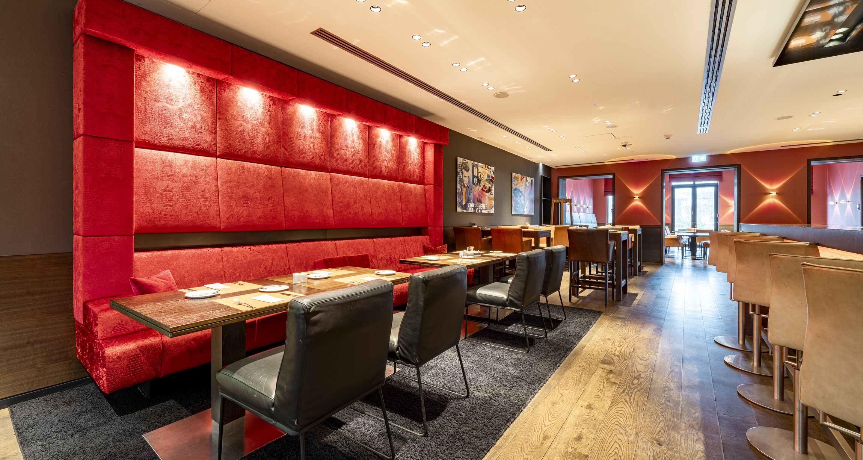 A modern restaurant with red upholstered booths, wooden tables, and black leather chairs.