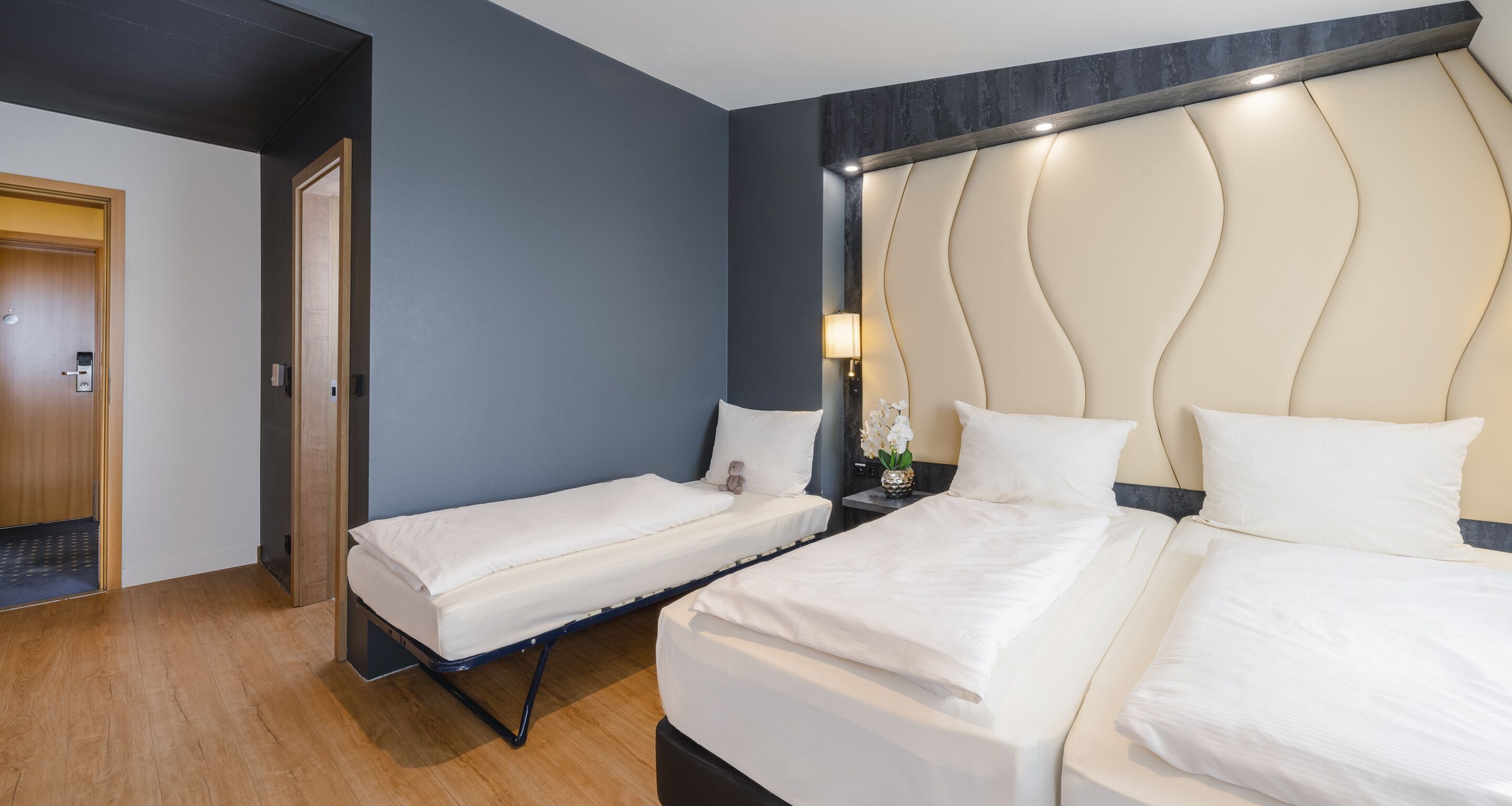 A hotel room with two single beds and a double bed, a wooden floor, a dark blue wall, and a marble headboard.
