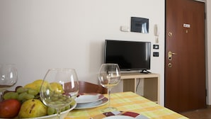 Un coin repas avec une table dressée pour deux personnes, un panier de fruits et un téléviseur posé sur un meuble.
