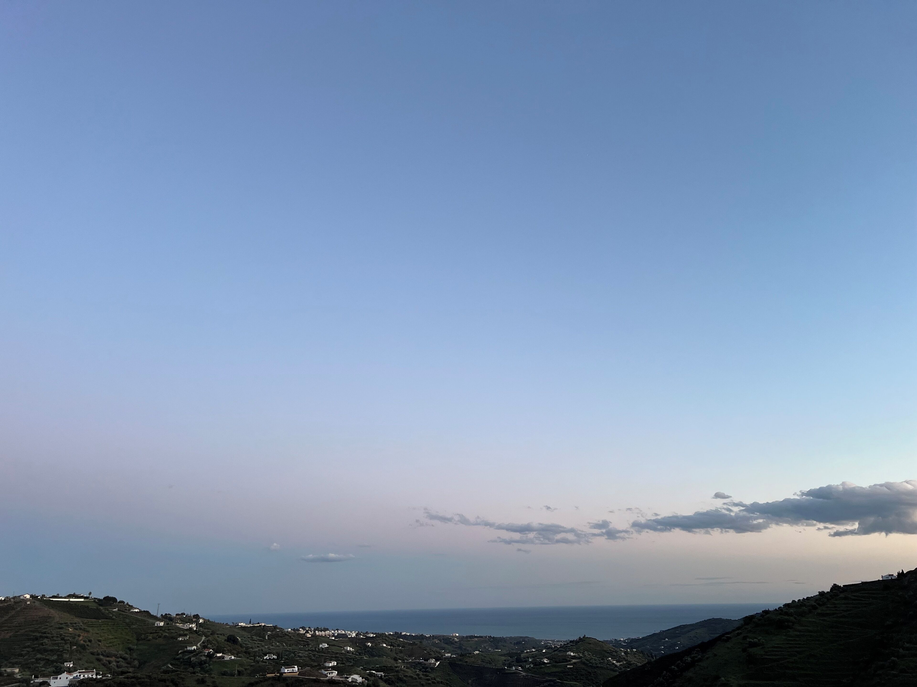 Paisagem costeira com céu claro, vista distante do mar e edifícios dispersos nas colinas.