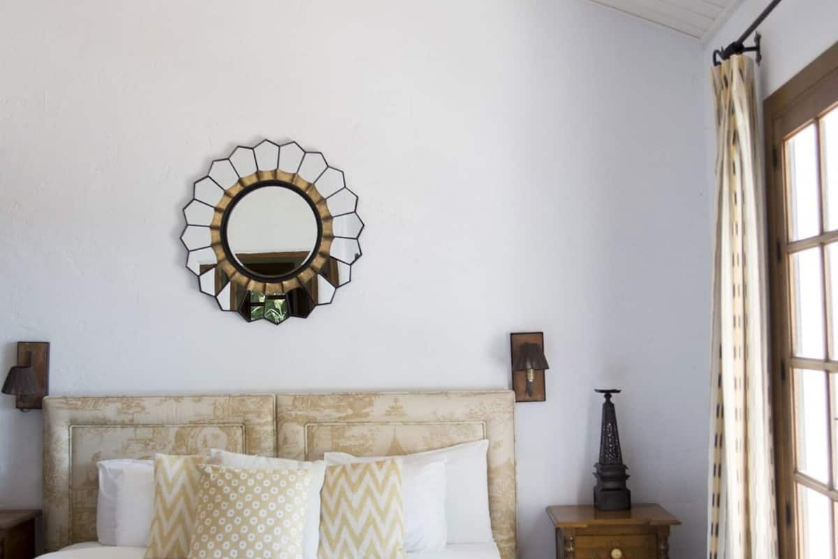 Un dormitorio con cama, un espejo redondo, una mesita de noche, un aparador y una ventana con cortinas.