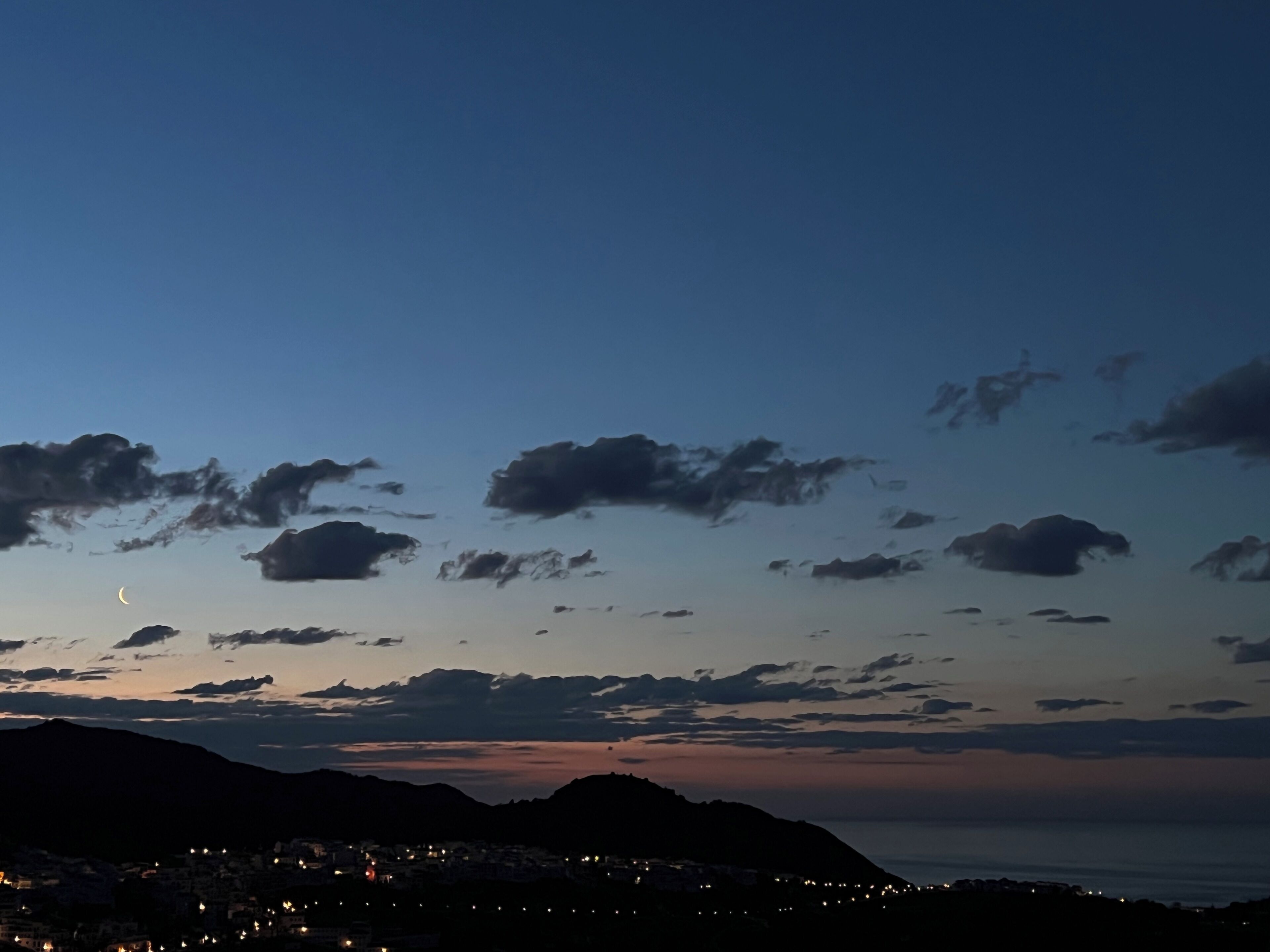 Un paisaje urbano costero al anochecer, con una luna creciente, nubes dispersas y montañas a lo lejos.
