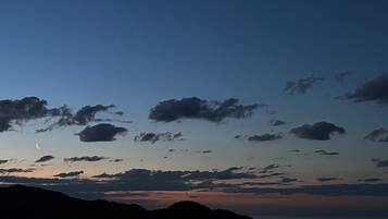 Un paisaje urbano costero al anochecer, con una luna creciente, nubes dispersas y montañas a lo lejos.