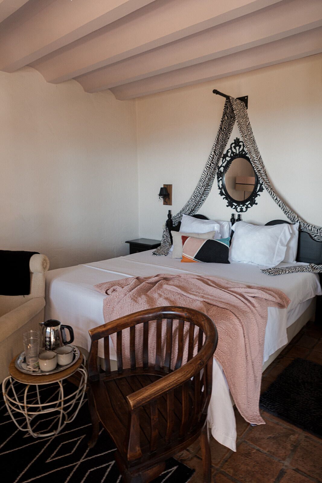 A bedroom with a canopy bed, a wooden chair, a bedside table with a kettle, and a small round table with a tray.