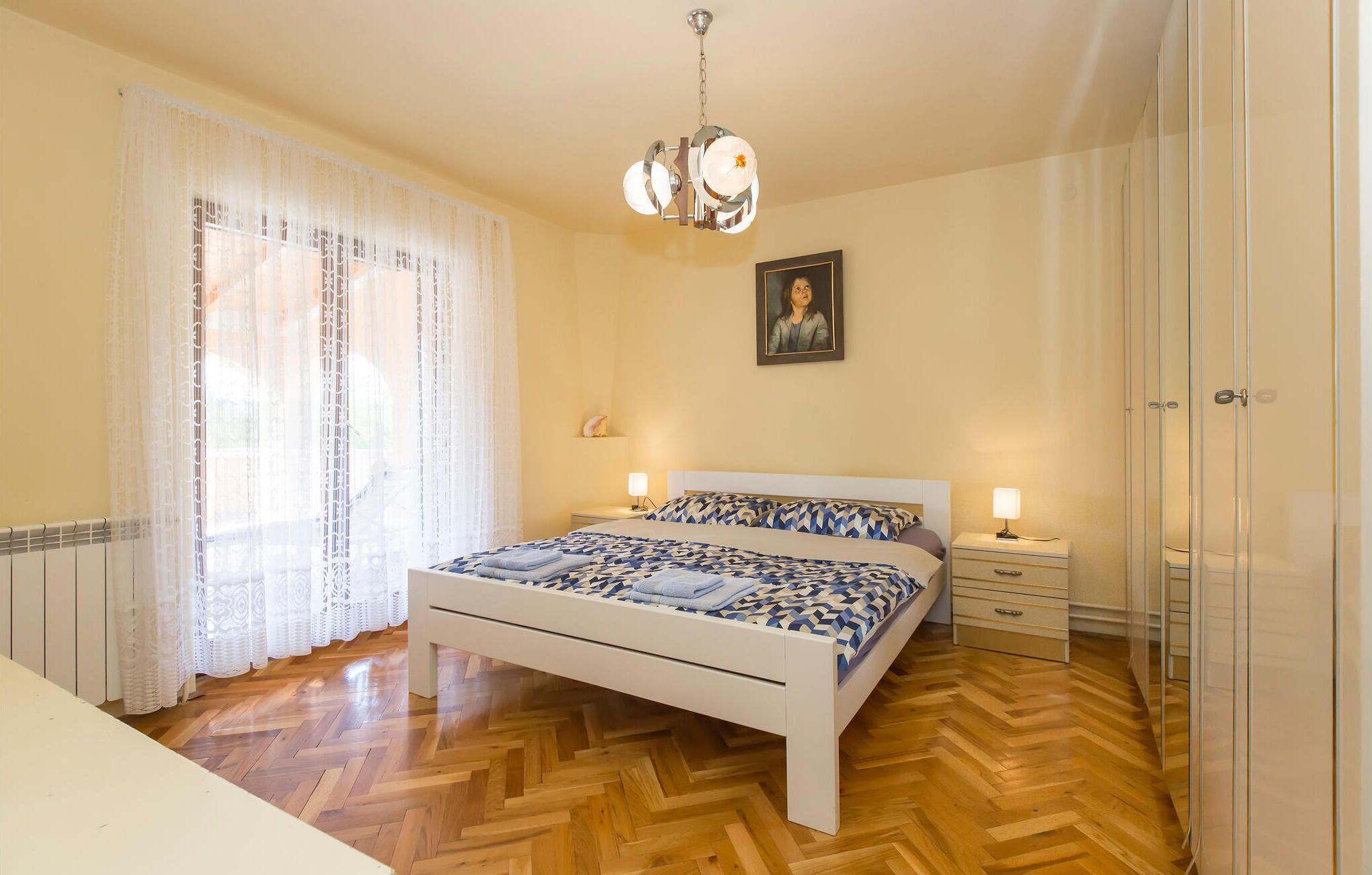 A bedroom with a bed, bedside tables, a chandelier, a radiator, and a window with curtains.