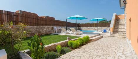 A backyard with a stone patio, a swimming pool, and two blue umbrellas.