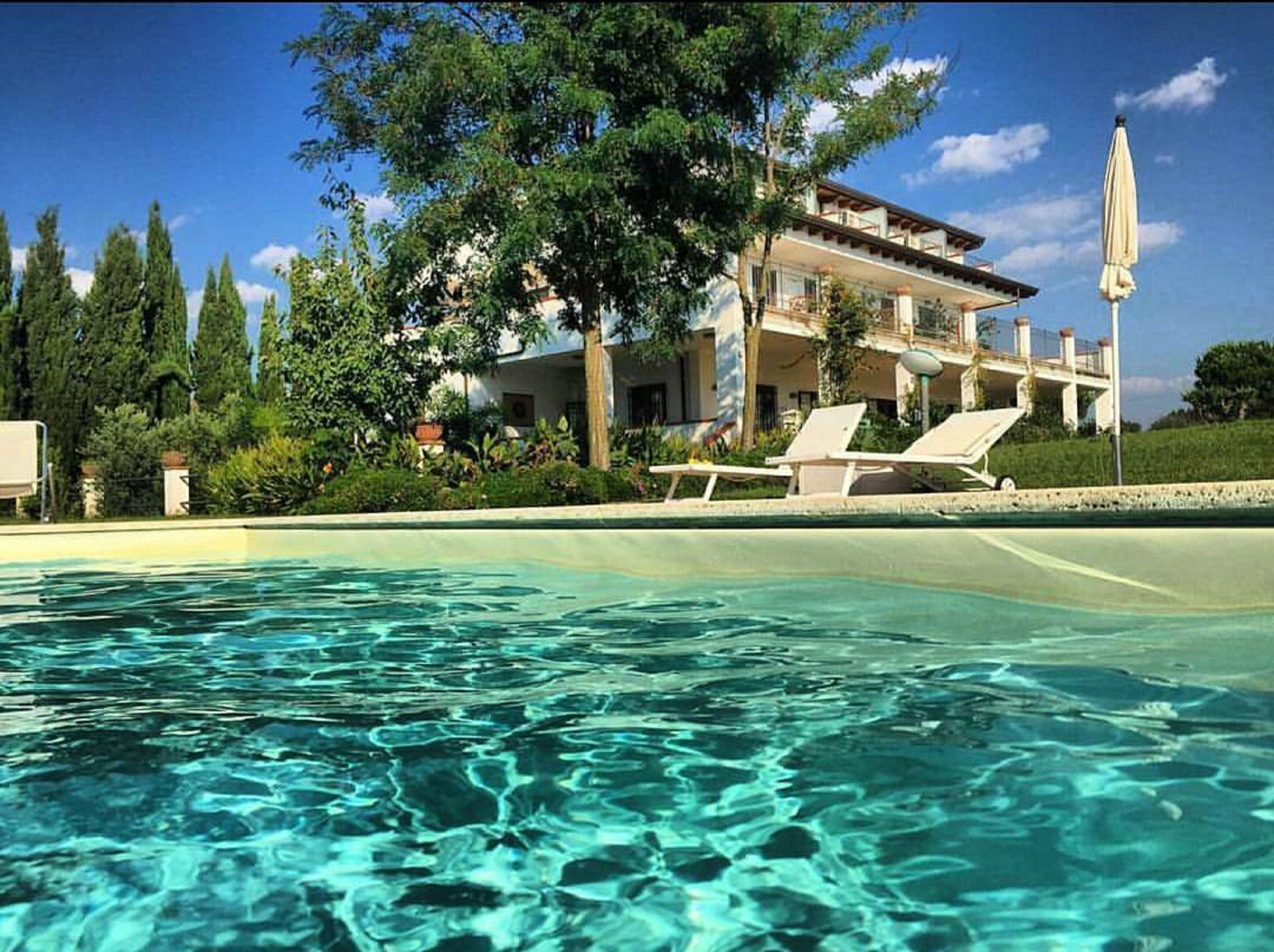 Una piscina con acqua cristallina, circondata da rigogliosa vegetazione e un grande albero. Si vedono lettini e, sullo sfondo, un edificio con un balcone.