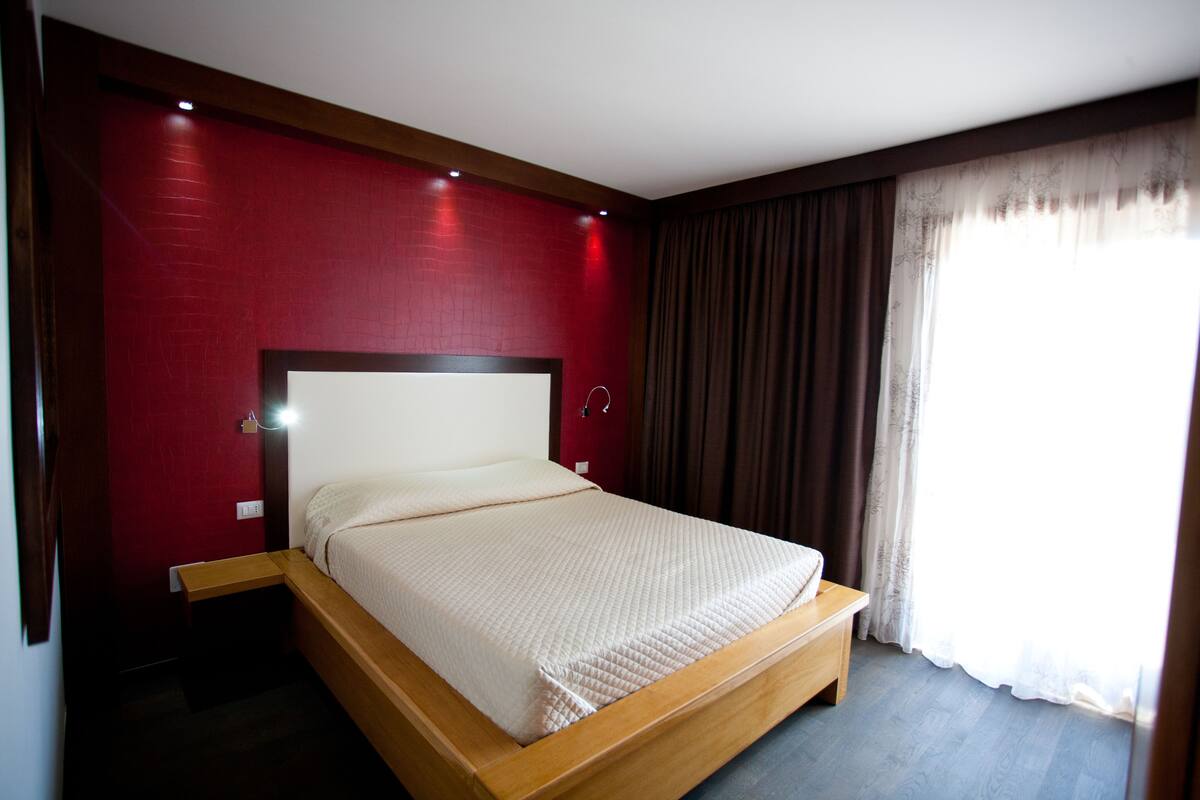 Una cama individual con estructura de madera y ropa de cama blanca en una habitación con paredes rojas y un gran ventanal con cortinas.
