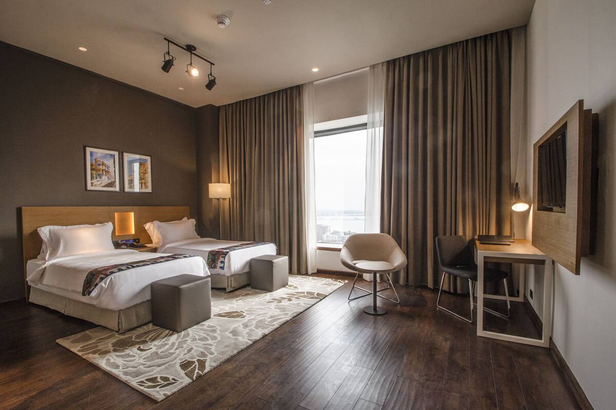Une chambre d’hôtel avec deux lits, un bureau, une chaise, une télévision et une grande fenêtre avec des rideaux.