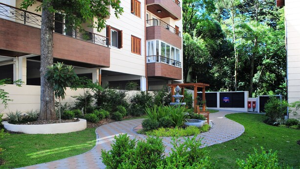 A multi-story residential building with a landscaped garden, a curved pathway, and a pergola. - Apartamento Flores da Montanha - 052 (Gramado)