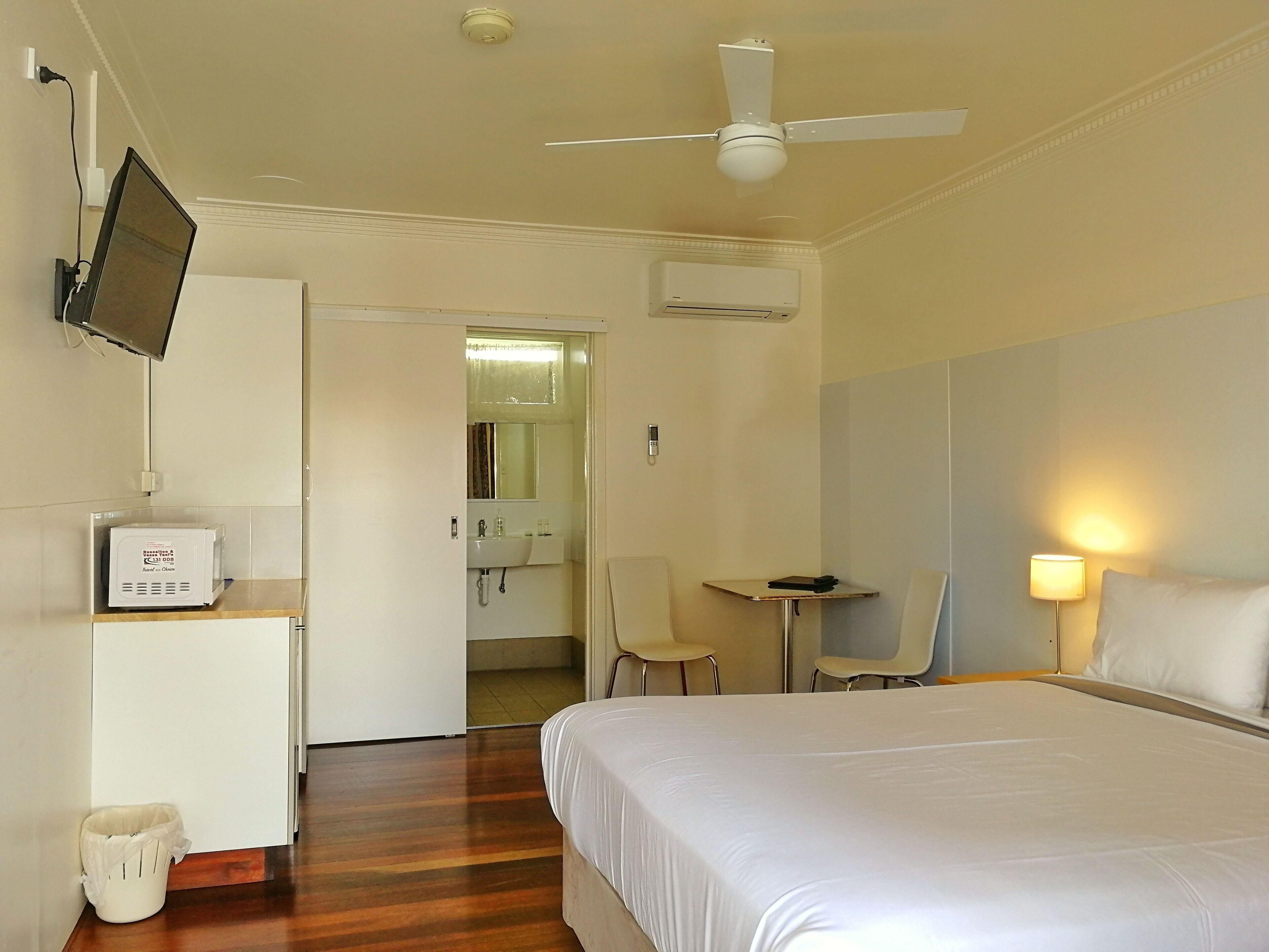 Een hotelkamer met een bed, een bureau, een stoel, een aan de muur bevestigde televisie, een airconditioning en een prullenbak.