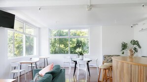 A bright, open-plan living area with a green sofa, white chairs, a wooden bar, and large windows offering a view of greenery. - Paradise Motel Busselton (Busselton)