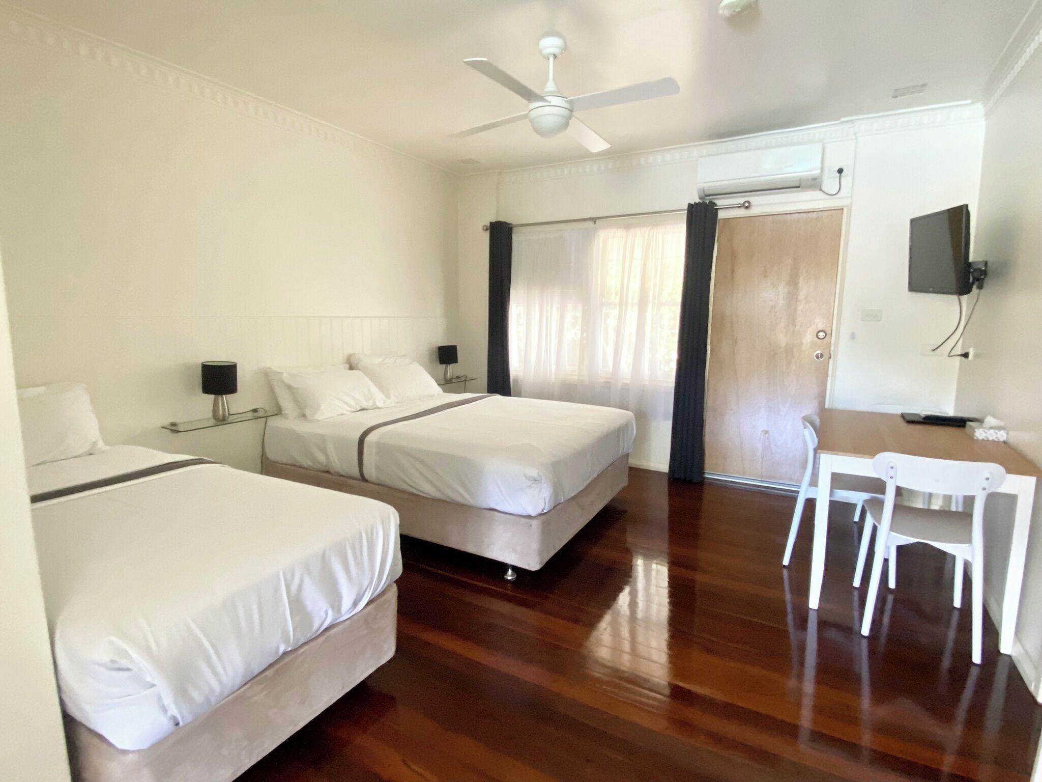 Habitación de hotel con dos camas, ventilador de techo, escritorio con silla y televisor montado en la pared.