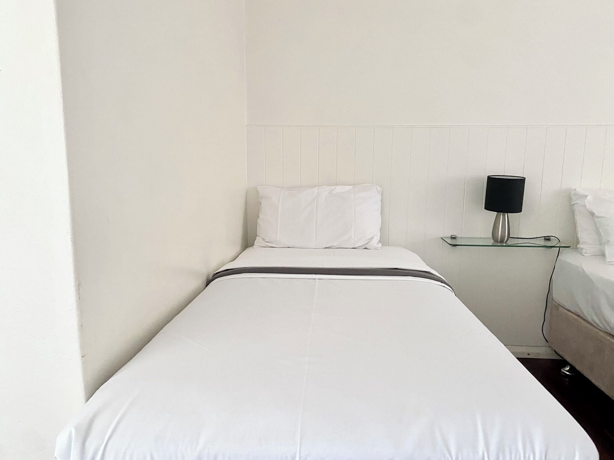 A small, single-bed room with a white bed, a bedside table with a lamp, and a white wall.