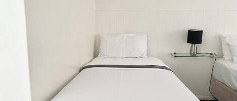 A small, single-bed room with a white bed, a bedside table with a lamp, and a white wall.