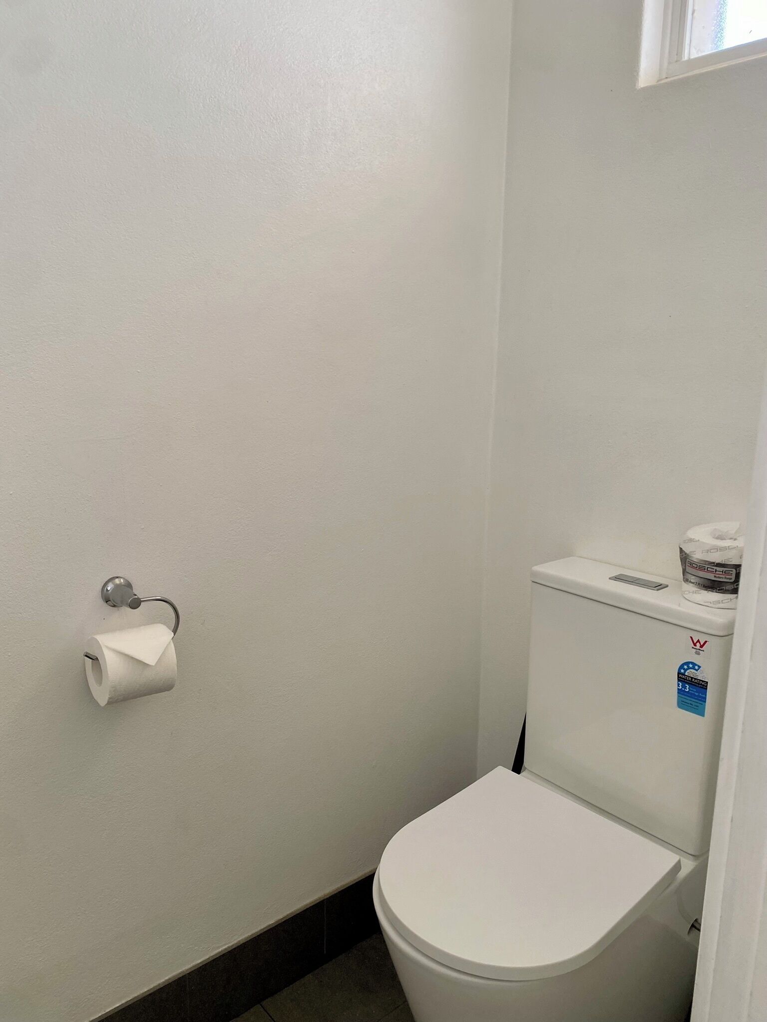 Et lille, hvidt toilet med tank og låg, toiletpapirholder på væggen, og et vindue over toilettet.