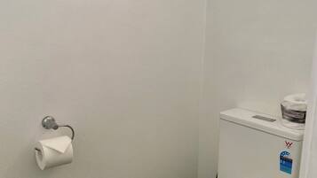 De petites toilettes blanches, avec un réservoir et un abattant, un porte-rouleau de papier toilette fixé au mur et une fenêtre située au-dessus des toilettes.