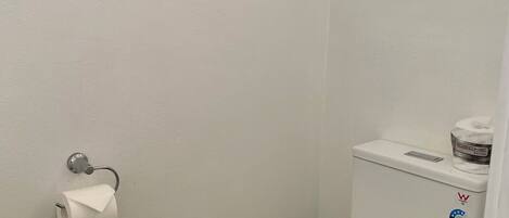 Un inodoro pequeño y blanco con tanque y tapa, un dispensador de papel higiénico en la pared, y una ventana sobre el inodoro.