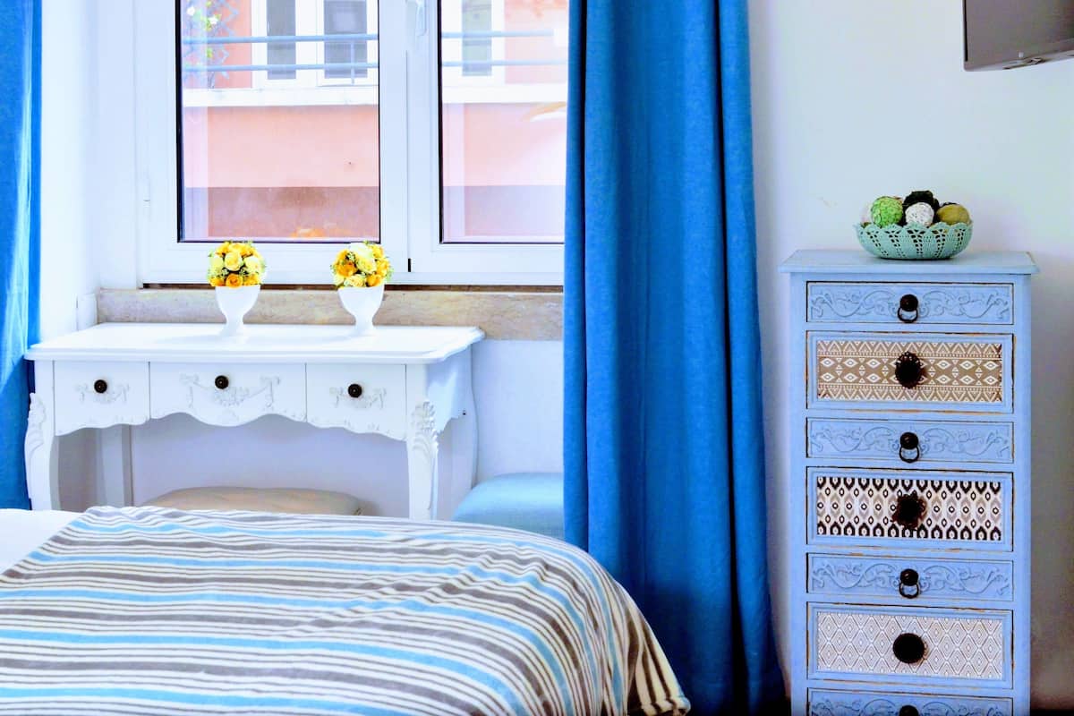 Une chambre à coucher avec un lit, une commode blanche aux tiroirs à motifs, une fenêtre avec des rideaux bleus et un saladier de fruits posé sur la commode.