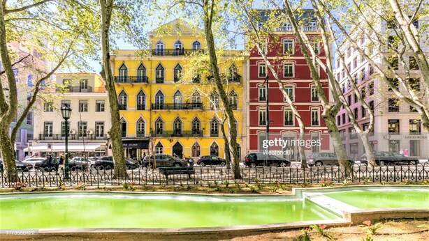 A street with colorful buildings, a green park area, and parked cars. - Guesthouse Sweet Liberdade (Lisbon)