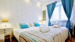 A neatly made bed with a striped blanket, two bedside lamps, a nightstand, and a window with curtains. - Guesthouse Sweet Liberdade (Lisbon)