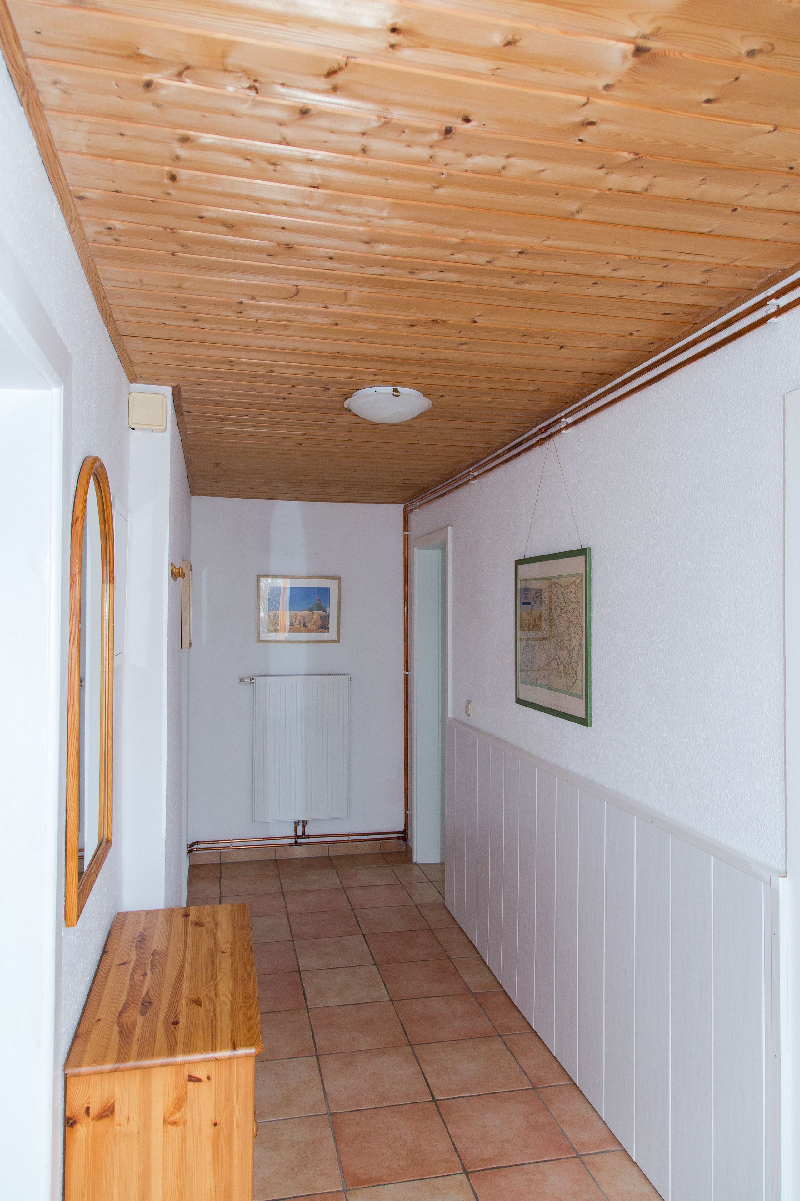 Un couloir avec un plafond en bois et des murs blancs, agrémenté d’un radiateur, d’un banc en bois et de tableaux encadrés accrochés aux murs.