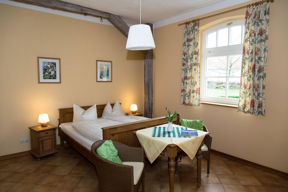 Une chambre à coucher avec un lit, des tables de chevet, une table recouverte d’une nappe à carreaux, une chaise et une fenêtre ornée de rideaux à motifs floraux.
