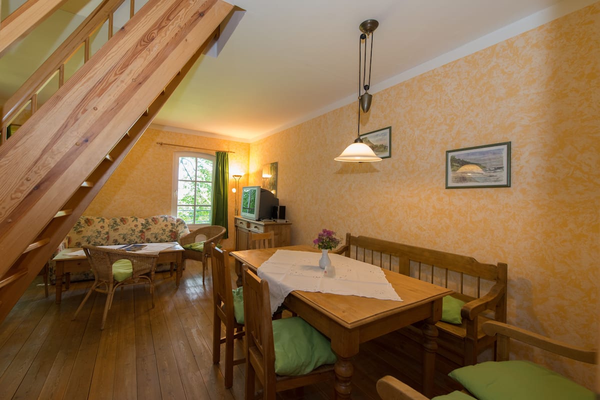 Un espace repas comprenant une table en bois, des chaises et un banc. Un escalier mène à un niveau supérieur.