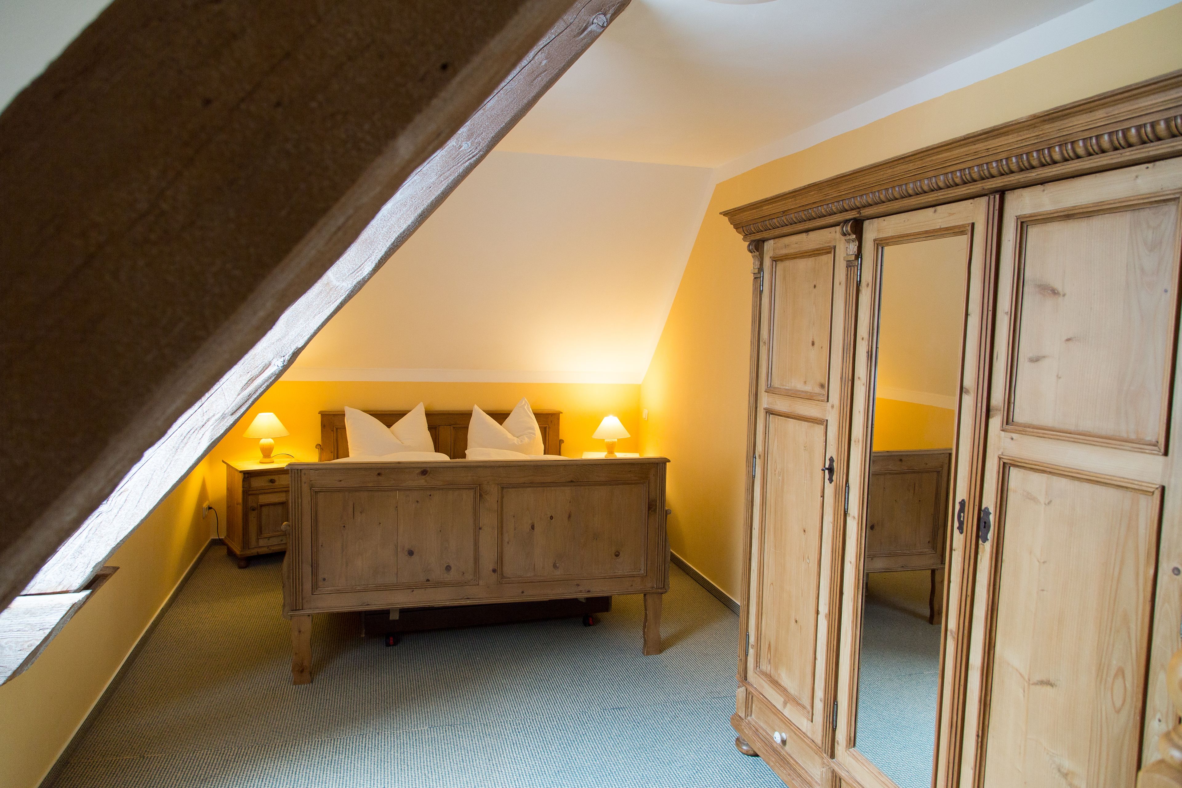 A bedroom with a sloped ceiling, a wooden bed with pillows, a nightstand with a lamp, and a wooden wardrobe.