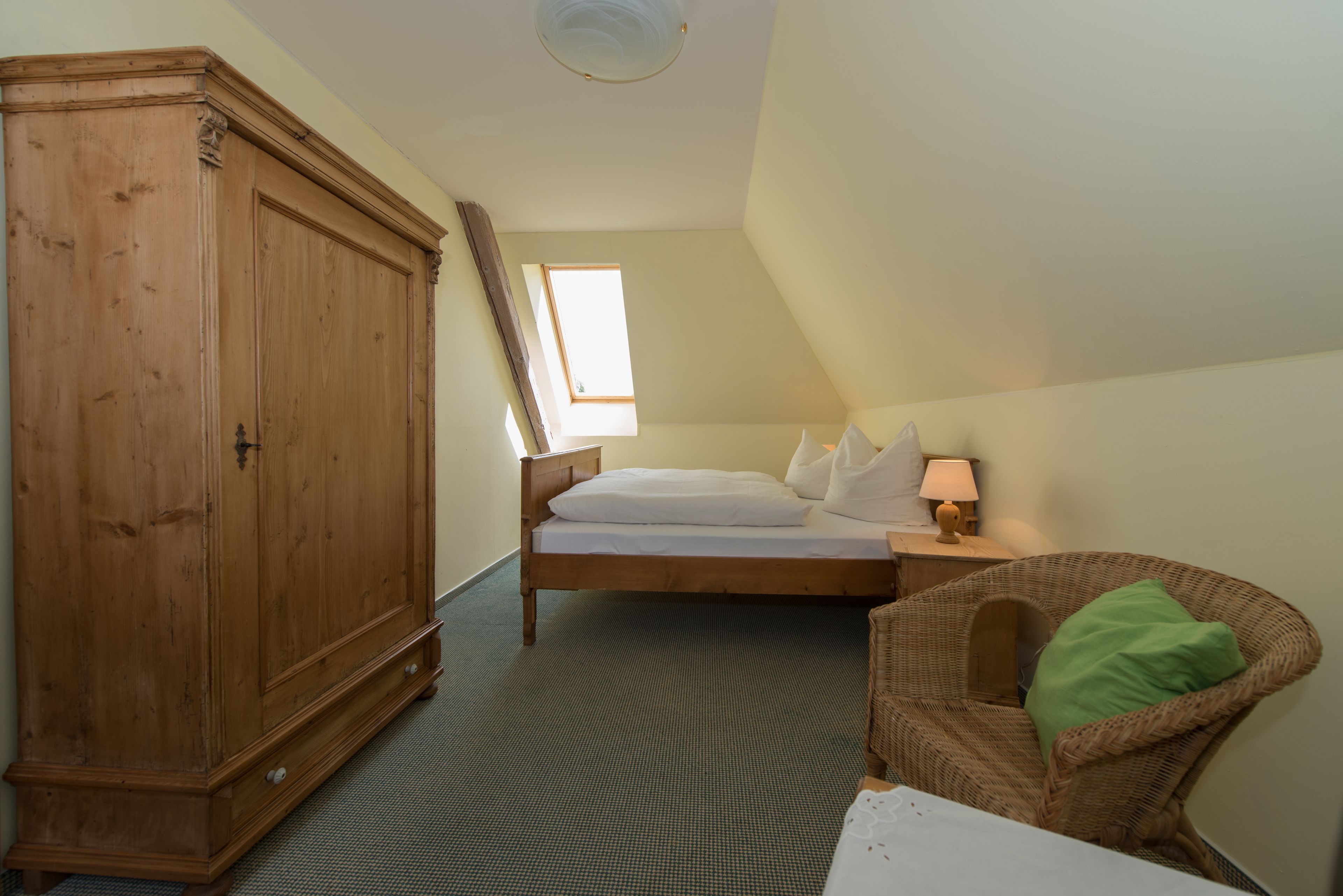 Une chambre avec une grande armoire en bois, un lit avec une literie blanche, un oreiller vert, une chaise en osier et une petite table de chevet avec une lampe.