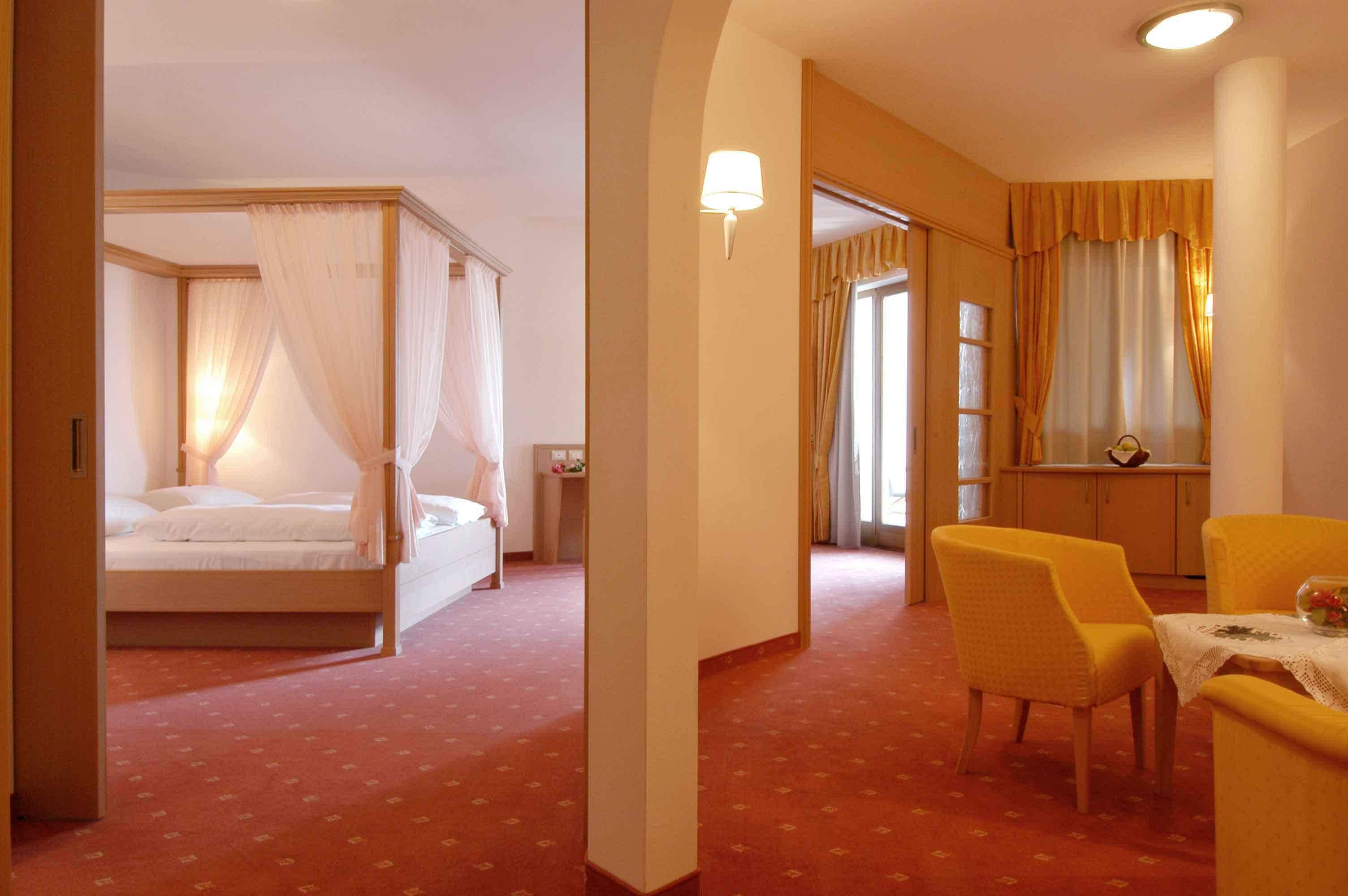 Ein Hotelzimmer mit Himmelbett, einer Sitzecke mit Tisch und Stühlen sowie Blick auf das angrenzende Zimmer.