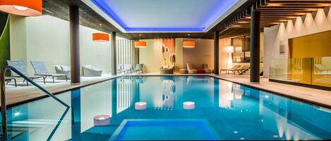 Una piscina interior moderna con agua cristalina, rodeada de sillas de descanso y elegantes lámparas colgantes naranjas.