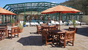 An outdoor seating area with wooden tables and chairs, potted plants, and a glass-enclosed pool area with lounge chairs. - Cardigan Bay Holiday Park (Cardigan)