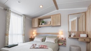 A neatly arranged bedroom with a bed, bedside tables, a mirror, and a small shelf with a plant and picture frame. - Cardigan Bay Holiday Park (Cardigan)