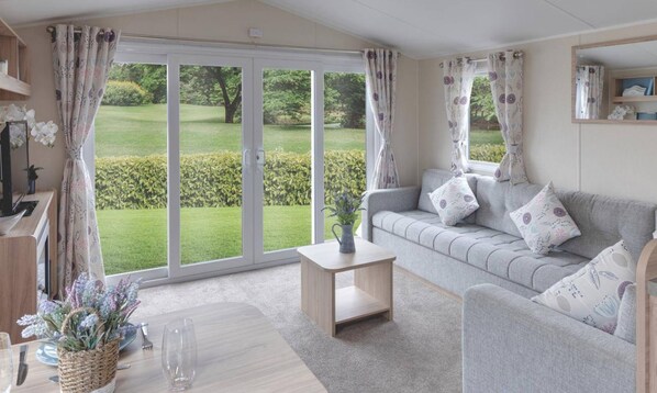 A cozy living room with a grey sofa, floral cushions, a small wooden coffee table, and a view of a well-maintained lawn through large sliding glass doors. - Cardigan Bay Holiday Park (Cardigan)