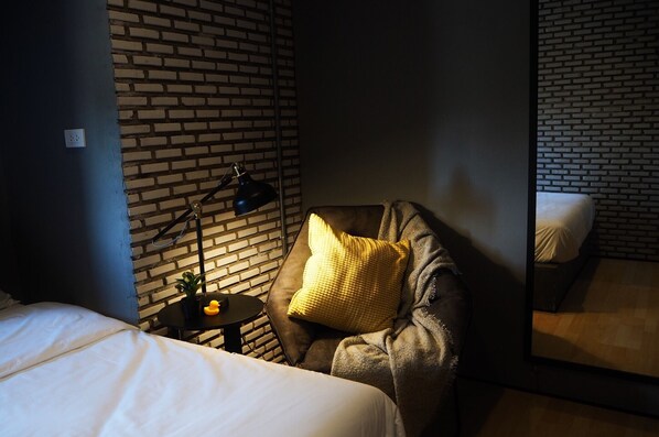 A hotel room with a brick wall, a bed, a chair with a yellow pillow, a small table with a lamp, and a mirror reflecting the room. - La Malila Hostel (Udon Thani)