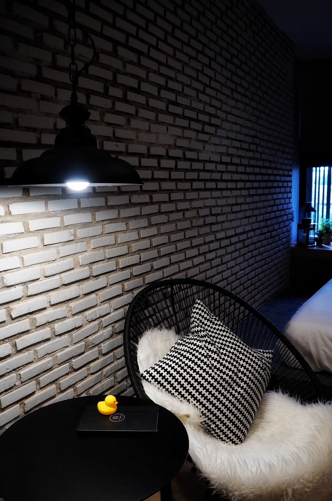 Una habitación con pared de ladrillo, una silla negra con cojín estampado, una pequeña mesa negra con un objeto amarillo y una lámpara colgante.