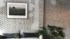Ein modernes Wohnzimmer mit einer weißen Ziegelwand, einem gerahmten Foto, einem grauen Sofa mit Kissen, einem runden schwarzen Tisch und zwei Zimmerpflanzen.