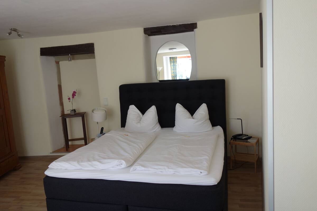 A neatly made bed with white linens in a room with wooden flooring, a wooden headboard, a small bedside table with a lamp, and a mirror on the wall.