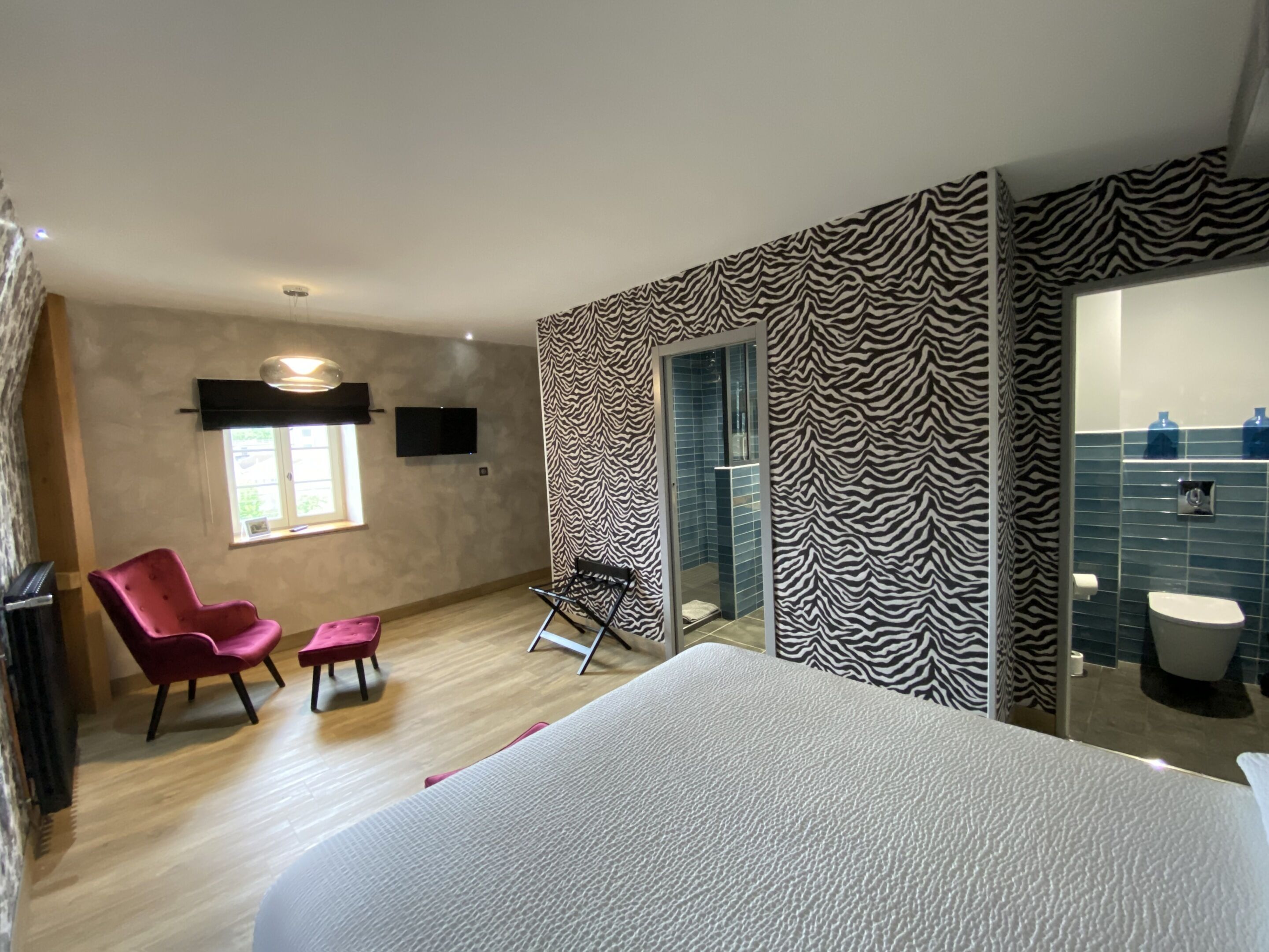 A hotel room with a bed, a red chair, a black folding chair, a zebra-patterned wall, a bathroom with a blue tiled wall, and a flat-screen TV mounted on the wall.
