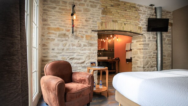 A hotel room with a stone wall, a bed, a chair, a small table with a water bottle, and a TV mounted on the wall. - Le Terminus (Tournus)